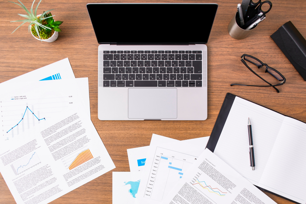 A view of a desk with a laptop and paperwork, including charts and a notepad, surround it.