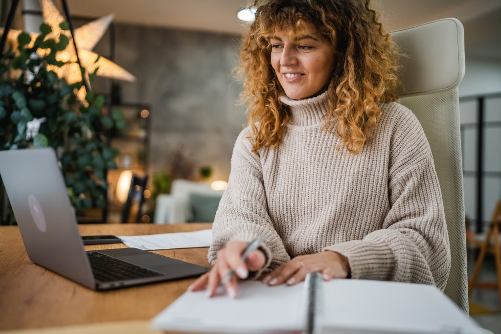 A woman is at her desk, in front of her laptop, with a notebook open in front of her, ready to take notes.