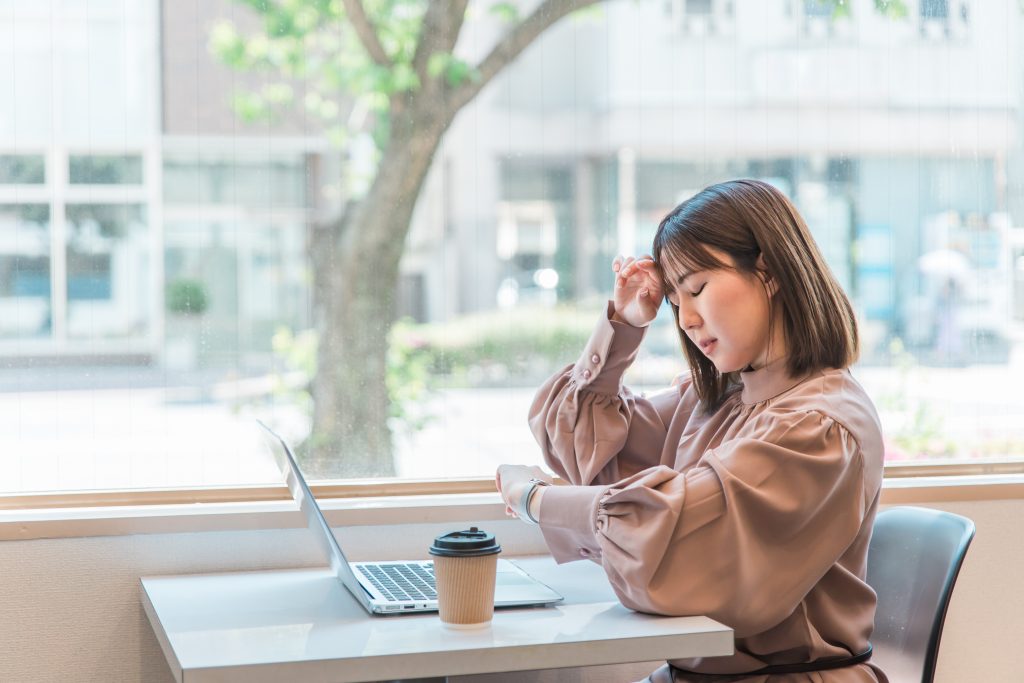 A woman is sitting at a table in a cafe with her laptop and a coffee, looking at her watch and feeling distressed that she missed a deadline.