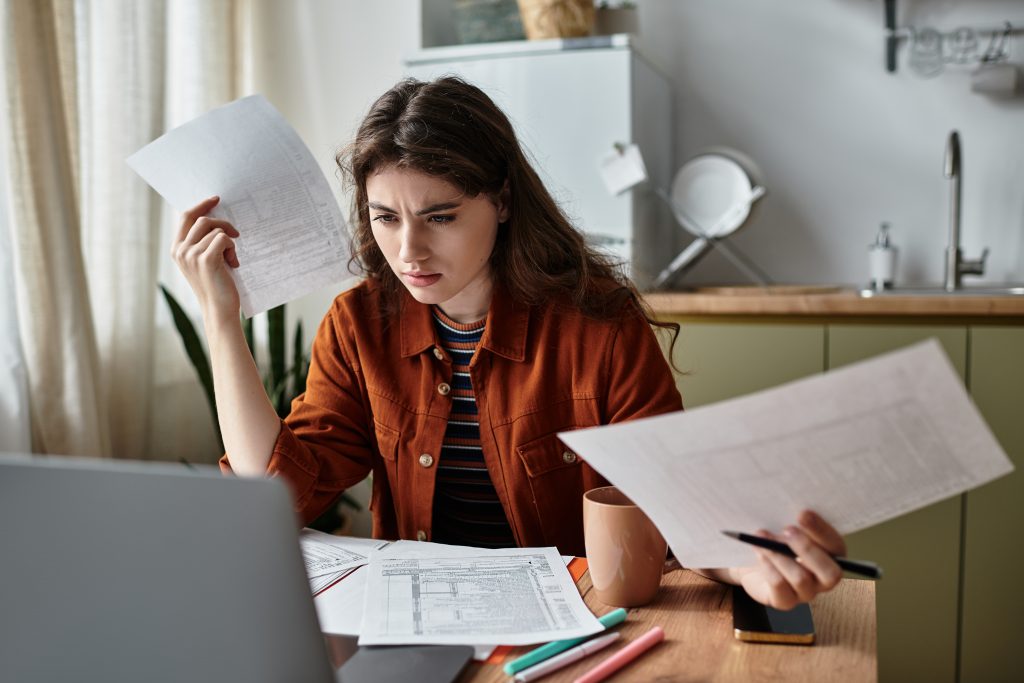 Woman at computer with multiple documents in her hands and on her desk, looking confused.