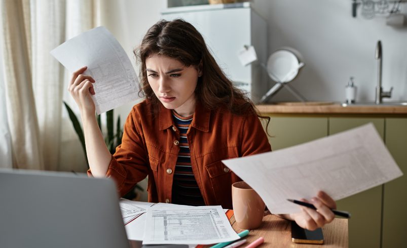 Woman at computer with multiple documents in her hands and on her desk, looking confused.